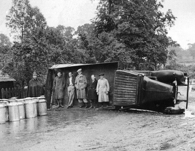 Passers-by use an overturned lorry as a shelter during a sudden shower of rain near Navestock, Romford, Essex. The lorry overturned on a sharp bend, discharging its load of milk churns, but the driver escaped uninjured.
15th June 1937. (Photo by Fox Photos)
